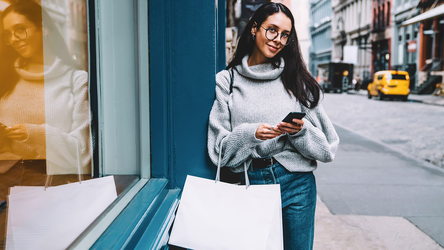 Young female standing on city street with shopping bags and mobile phone