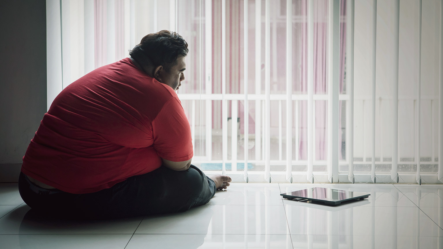 Obese man sitting on the floor looking out the window