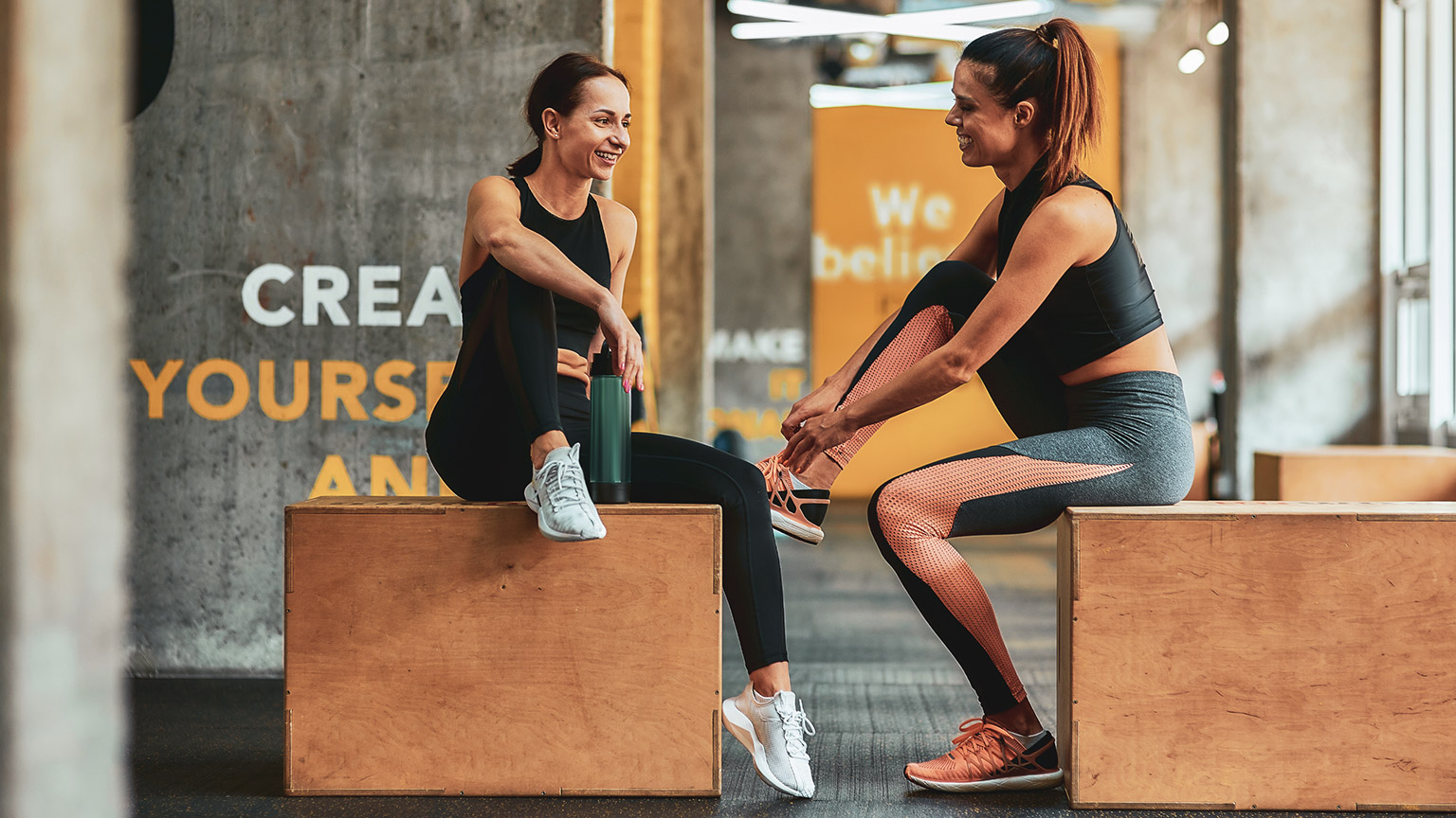 Two women talking while sitting on boxes in a gym