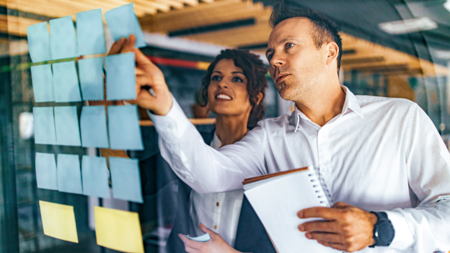 Male and female coworkers with serious faces pointing at sticky notes on a glass window