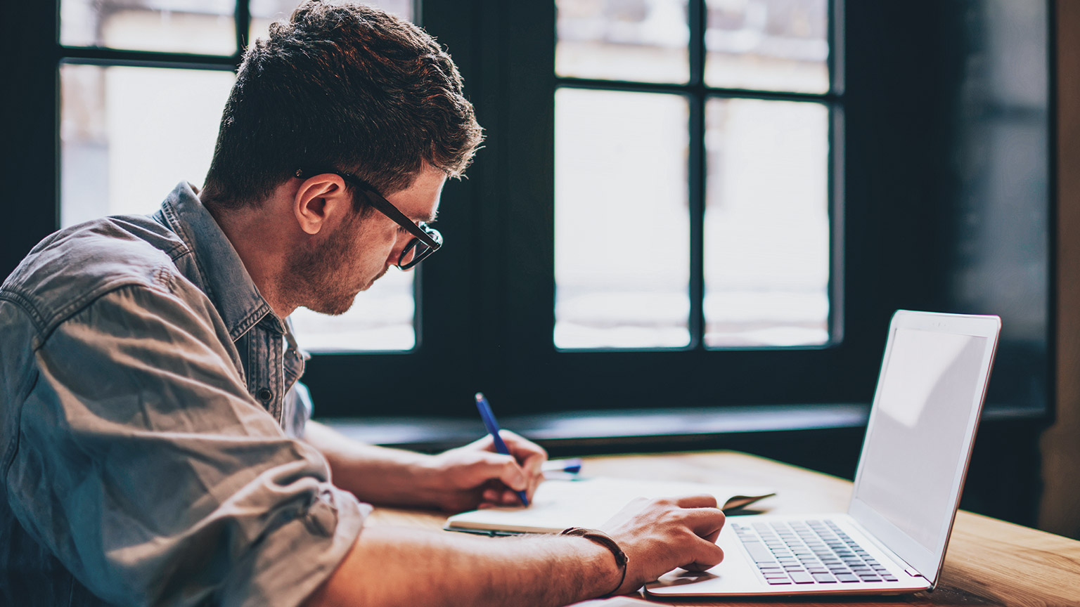 Side view of young male hipster sitting at a desk writing on a notepad as he prepares to use some of the course templates on his laptop