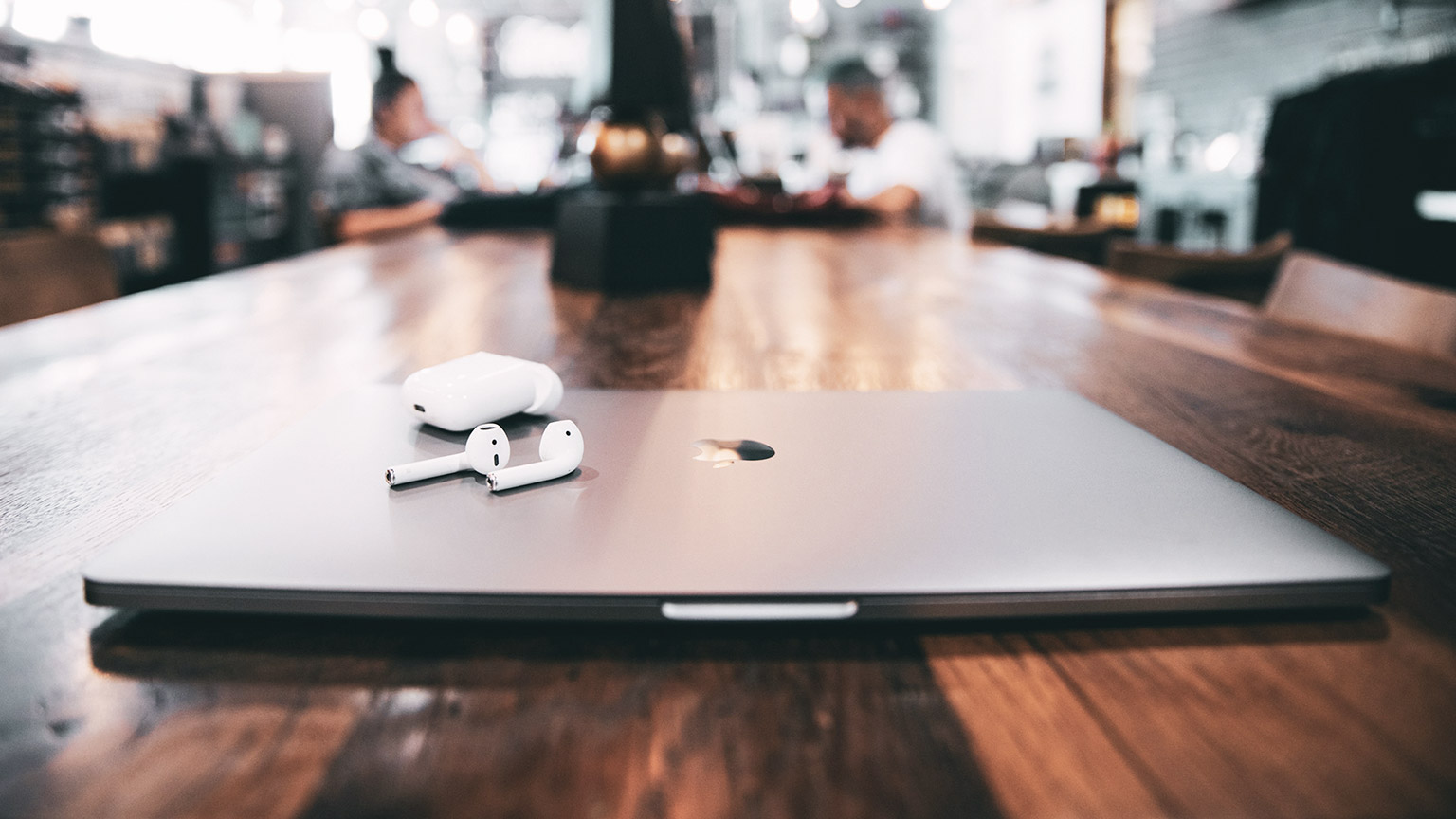 A shallow focus view of a laptop and airpods sitting on a desk with people working in the background