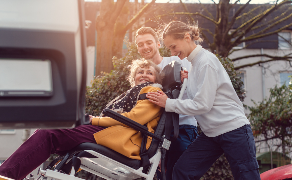 People wheeling elderly woman in her wheelchair into a van