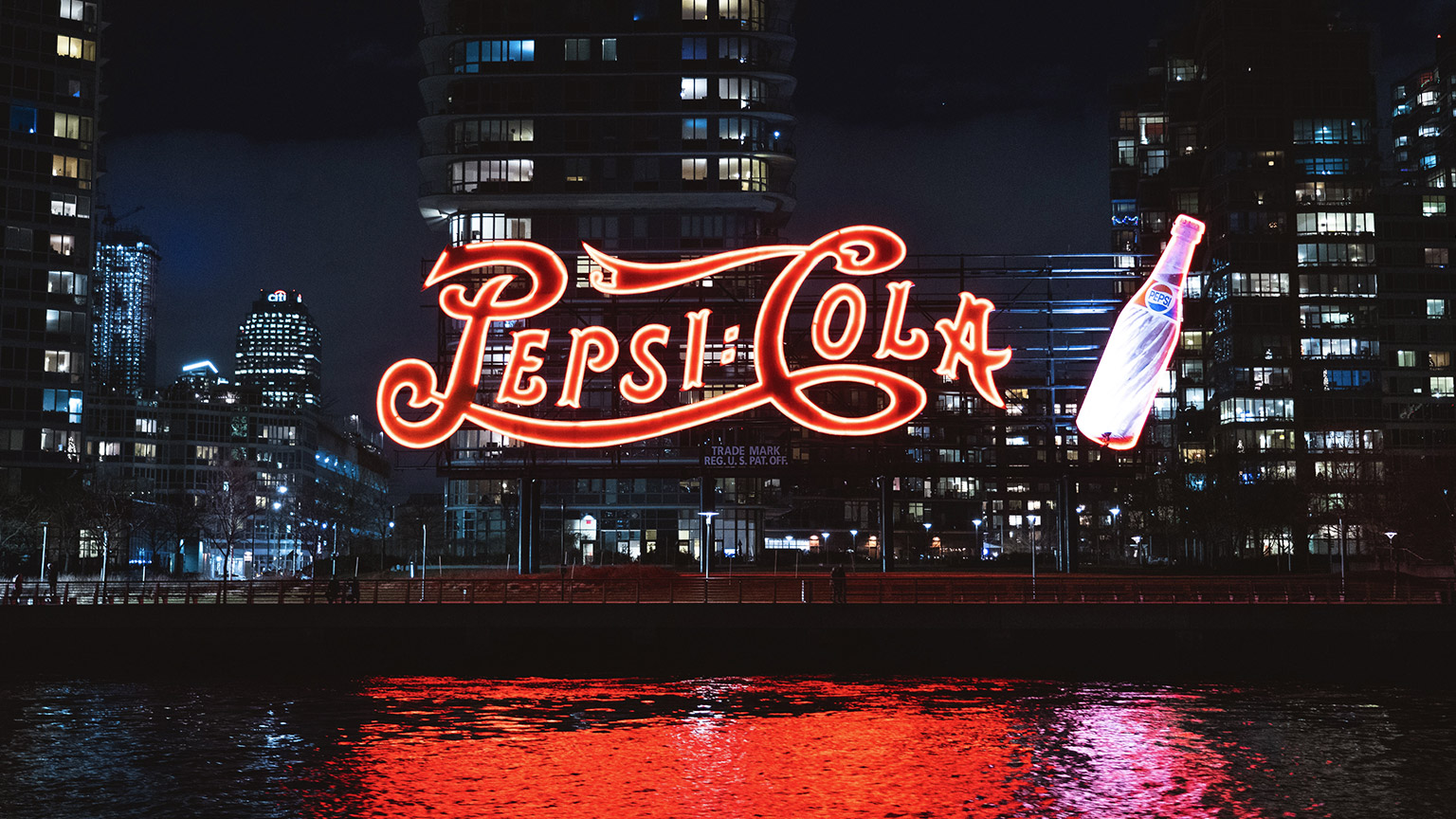 A neon pepsi sign by the side of a river in a city