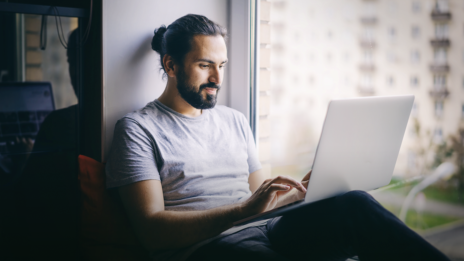 young dark-haired man working on laptop at home, sitting on windowsill with city view