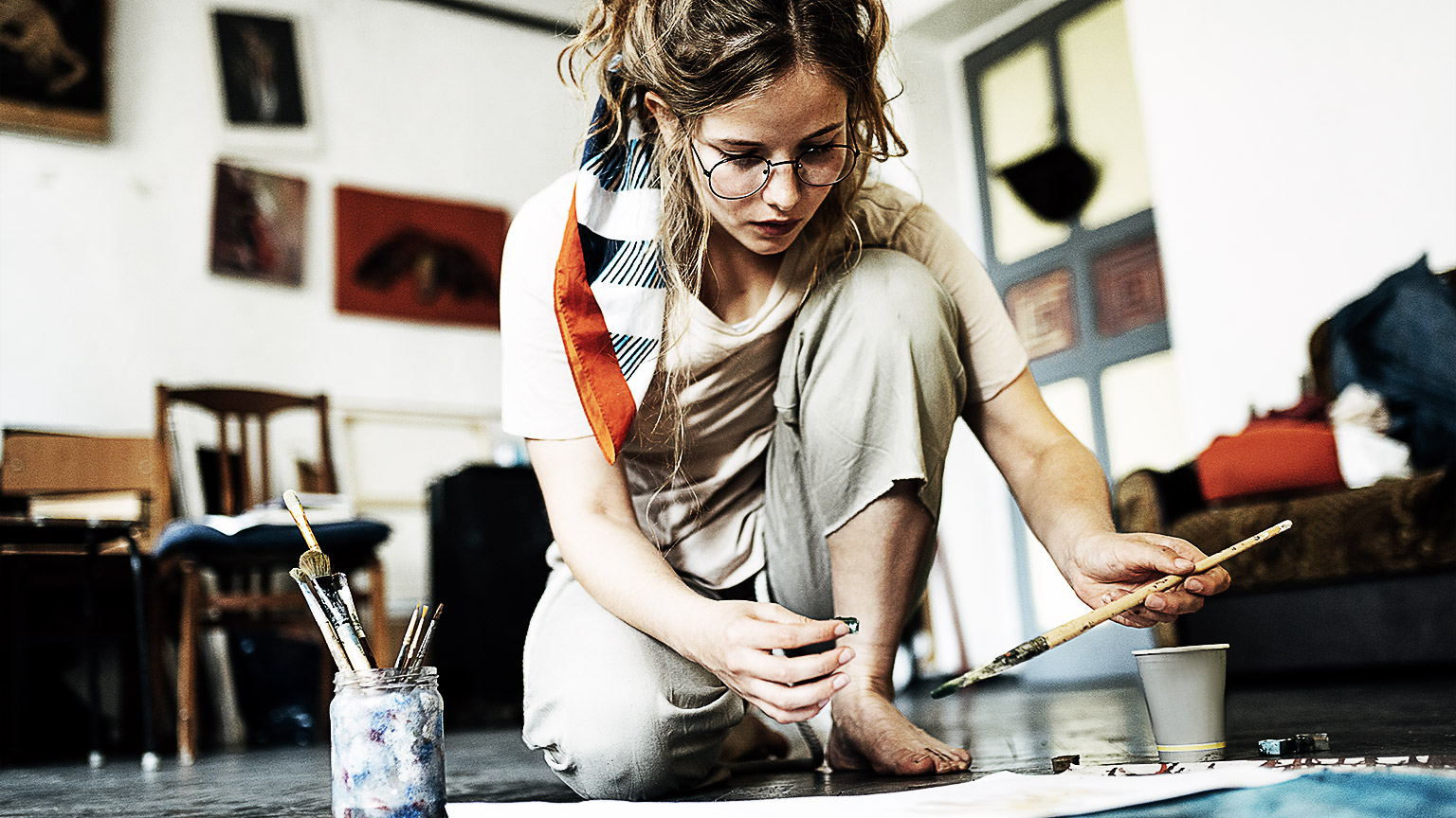 Horizontal image of a female artist sitting on the floor in the art studio and painting on paper with a brush.