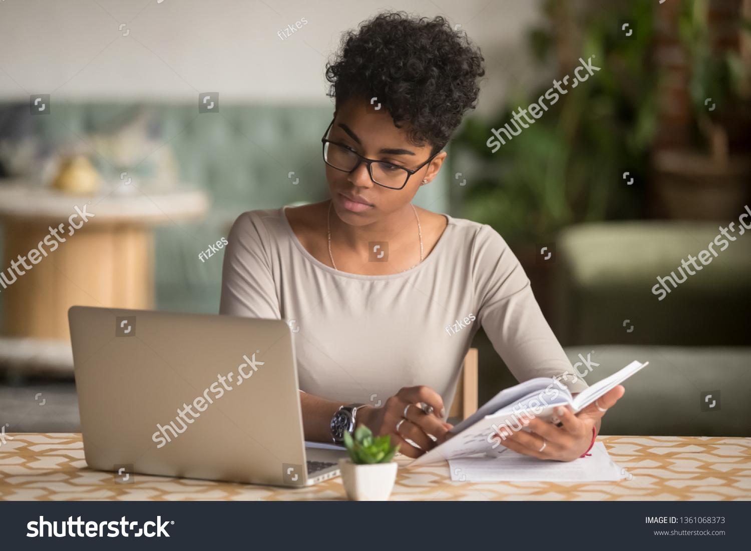 Focused young african american businesswoman or student looking at laptop holding book learning, serious black woman working or studying with computer doing research or preparing for exam online