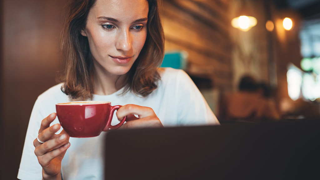 female manager holding in hands cup of coffee looking screen laptop in cafe