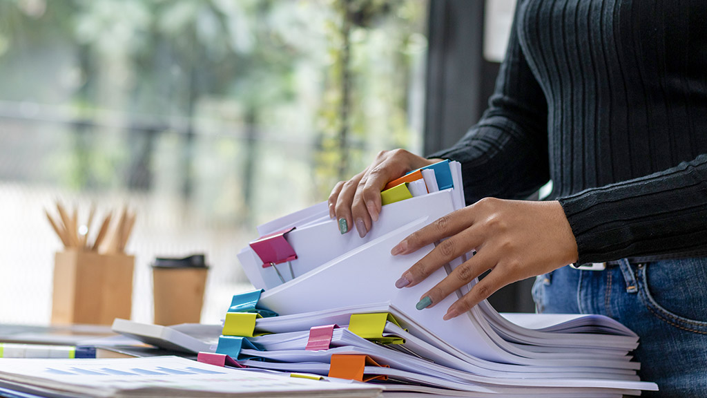 secretary searches through stacked documents on desk in office