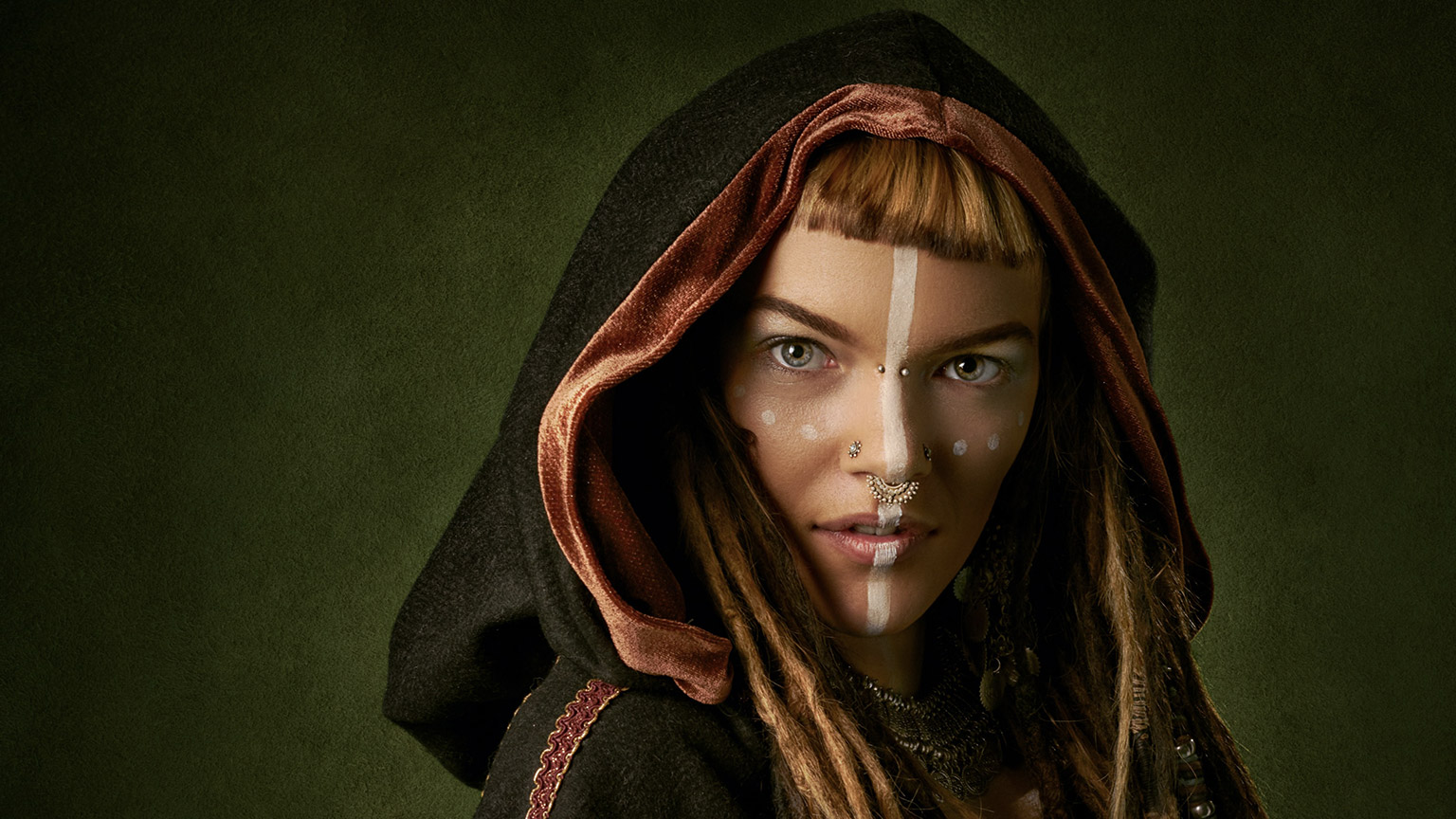 An female character with a white line down the middle of her face, wearing a cloak and many facial piercings. 