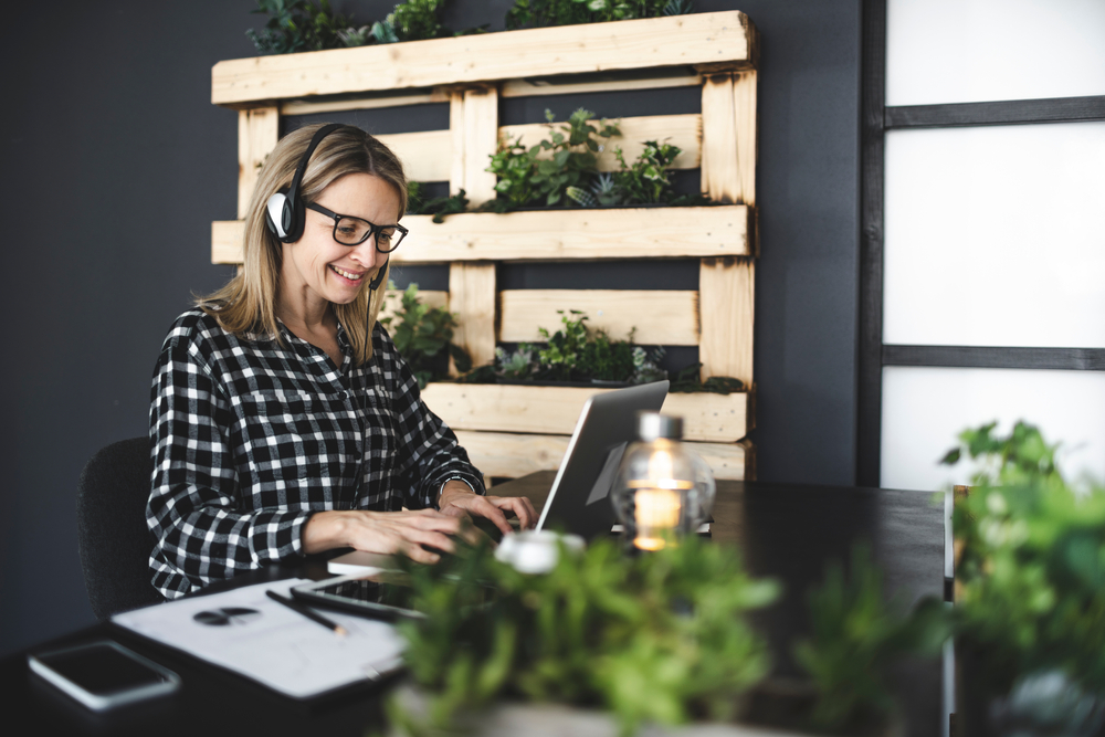 Female entrepreneur wearing black and white checked shirt is sitting in a sustainable, ecological office with a headset and is having an online meeting.