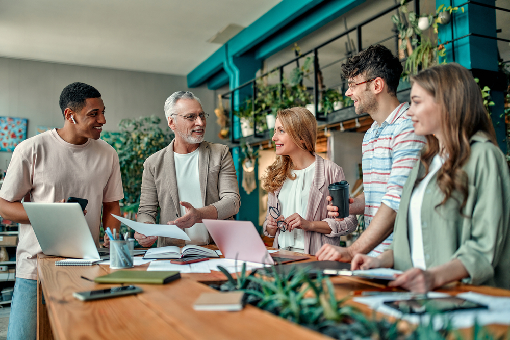 Group of young business people and senior team leader are working together with laptop around a wooden table with office plants