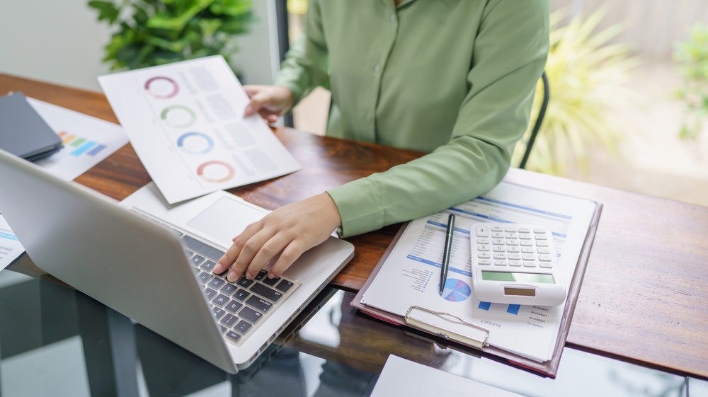 Accountant working in modern working space on a laptop, at wooden and glass desk, indoor plants behind her as she is reviewing financial reports.