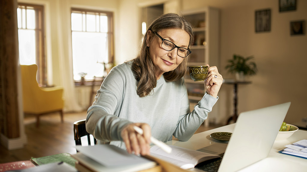 woman working from home with cup of coffee sitting at desk with books and laptop