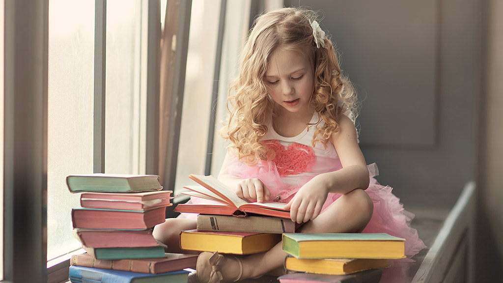 Cute curly little girl in a pink skirt reading a book, sitting on the window