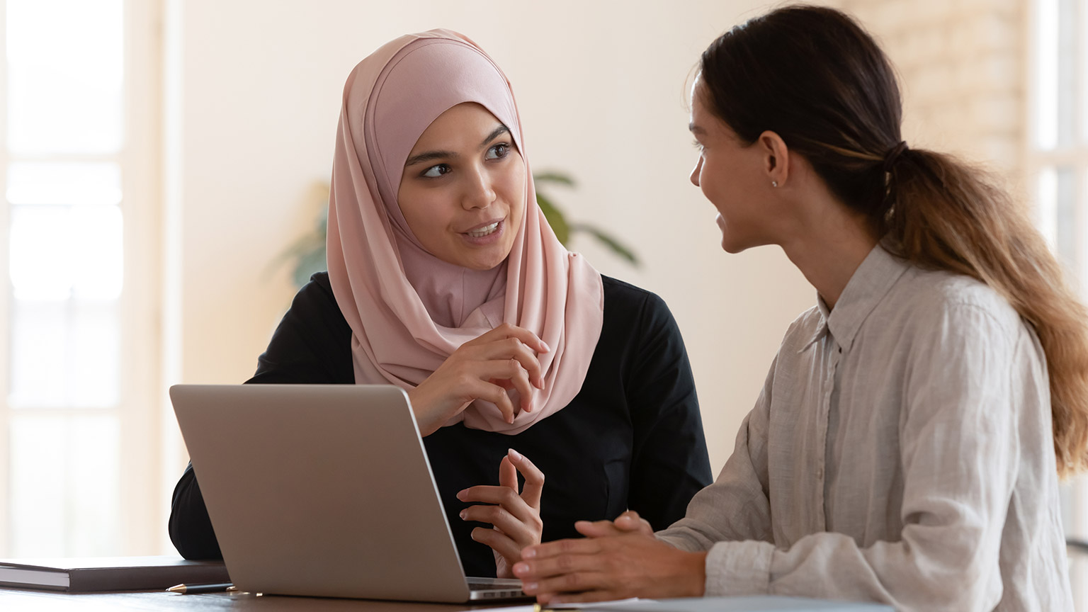 Multiracial young female employees work on laptop discuss business cooperation ideas at meeting together, international businesswomen brainstorm collaborating using computer at office briefing