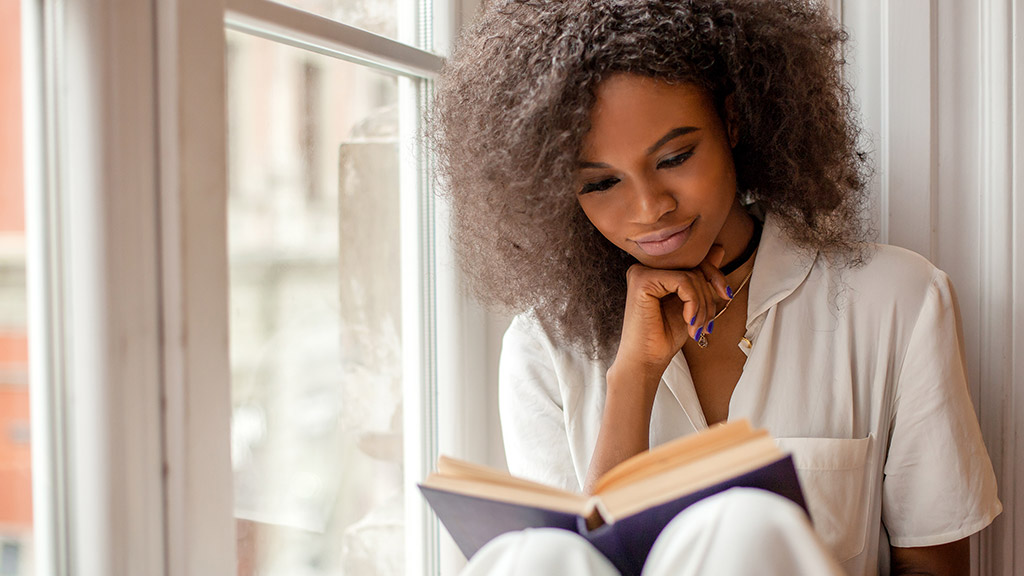 Pretty african american girl with reading a book sitting on the windowsill.