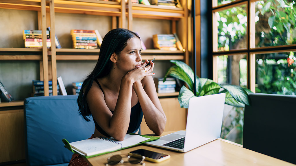 young female sitting at table brainstorming with netbook and opened copybook in light workspace