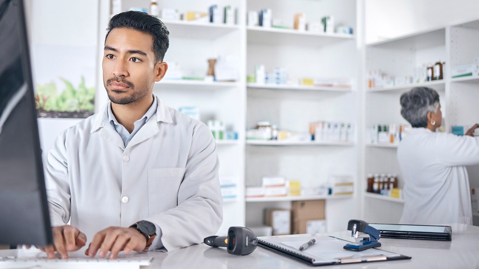 Pharmacist in foreground types at a desktop. A colleague works to stock the shelves behind.