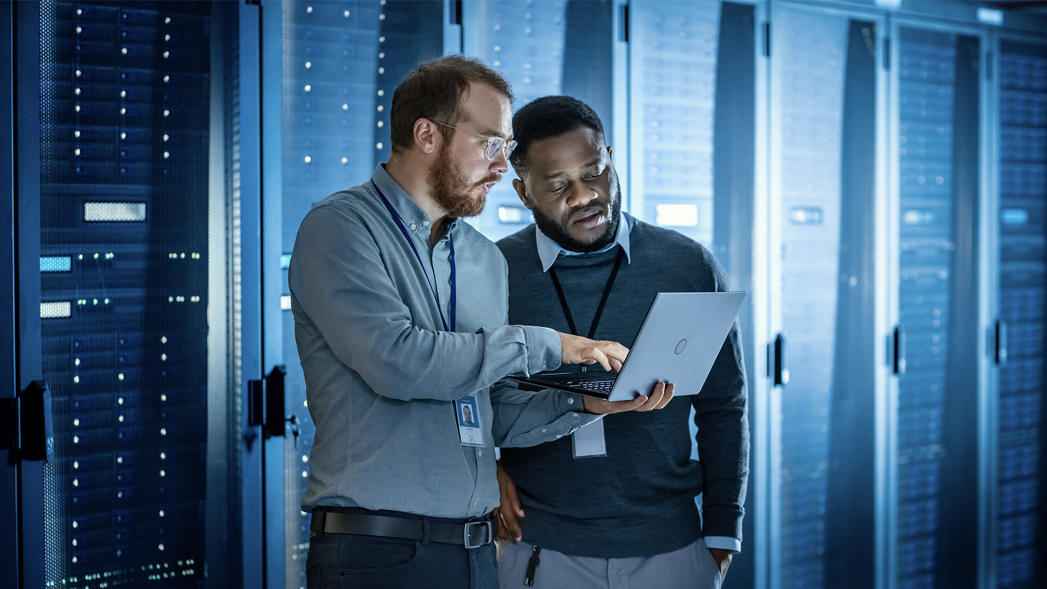 Male Engineer Colleague are Using Laptop in Data Center while Working Next to Server Racks Running Diagnostics or Doing Maintenance Work