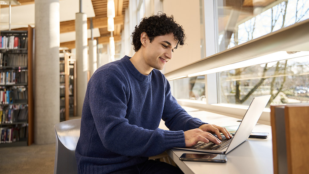 Smart Hispanic male student sitting at desk and using laptop