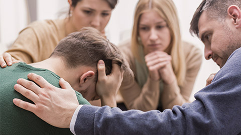 Close-up of a devastated young man holding his head in his hands and friends supporting him during group therapy