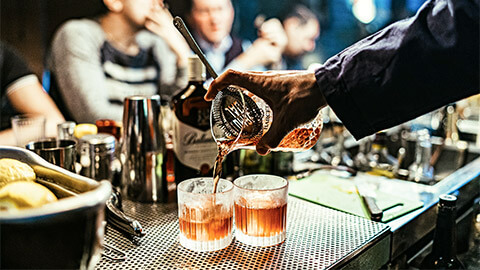 A bartender pouring cocktails into chilled tumblers at a busy cocktail bar