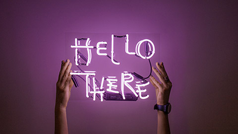 A person holding up an illuminated, neon sign that reads 'Hello there'