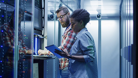 man and woman with tablet using laptop in server room while checking servers