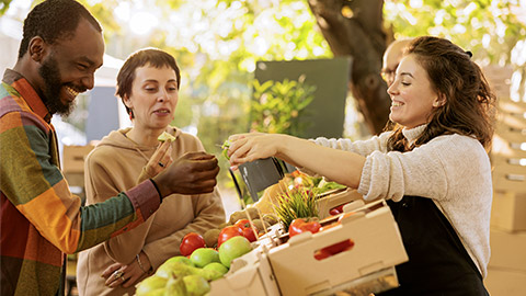 Happy multicultural couple tasting bio organic produce at farmers market, buying locally grown fruits and vegetables.