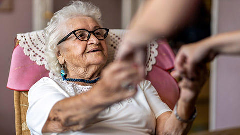 Female home carer supporting old woman to stand up from the armchair at care home