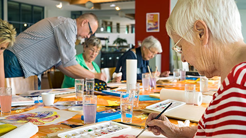 Woman in striped red and white shirt working on canvas while painting with brush at table with other students and teacher in spacious studio