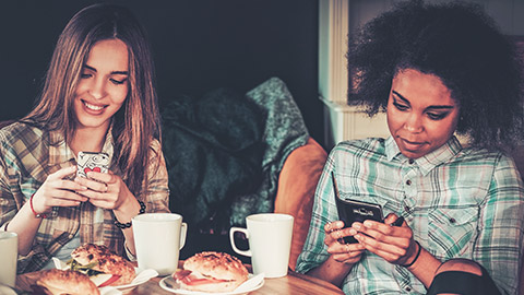 2 young females sitting in a cafe ignoring their food and focussing on their phones