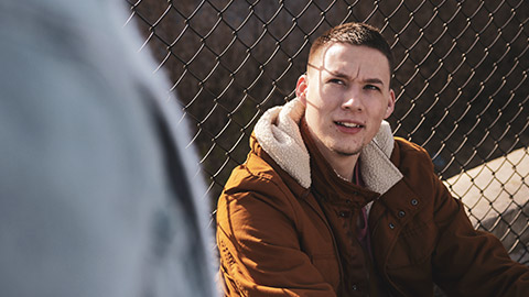 A teenager sitting on the ground near a fence looking up with a serious face at someone off camera