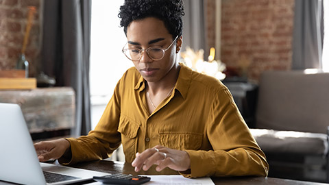 Focused millennial Black business woman calculating finance, money, using calculator, laptop computer at home workplace table