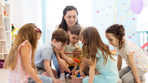 Preschool teacher plays with group of children sitting on a floor at kindergarten