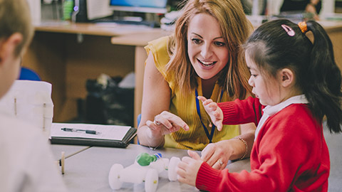 Teacher is sitting at the table in her classroom with her primary school students