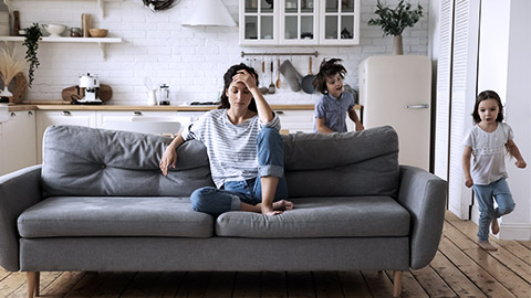 Woman looks tired sitting on couch with children playing