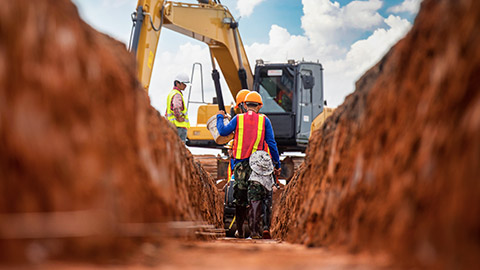 A low angle view of two workers in trench with an excavator in the background