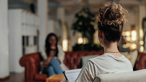 Focus on female psychiatrist writing information of african female patient while sitting on sofa and talking about his illness