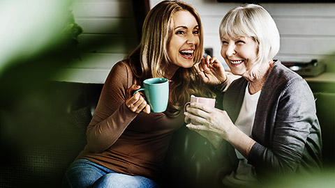 Happy senior mother with adult daughter sitting on couch and holding cups with coffee or tea at home