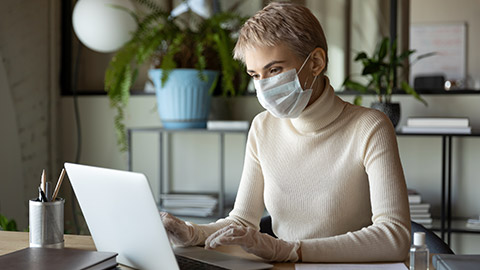 Concentrated young female employee worker wearing medical facial mask and protective gloves, using laptop in office