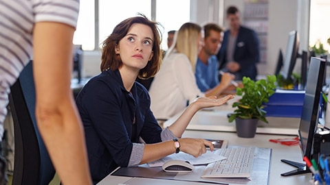 Young businesswoman in a serious discussion with a colleague gesturing towards her computer with her hand in a busy open plan office