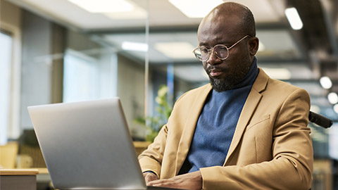 Bearded African American employee with laptop on his knees sitting in wheelchair and looking at screen while networking in office