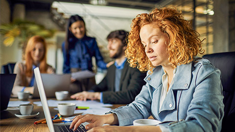 Focused businesswoman working on laptop with colleagues in background at office