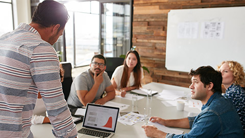 Young man giving business presentation on laptop to colleagues sitting around table in conference room