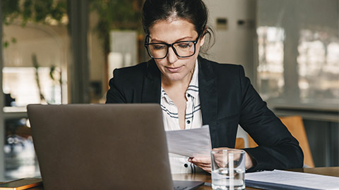 Photo of european businesswoman 30s wearing formal clothing and eyeglasses working in office on laptop and examining paper documents while holding