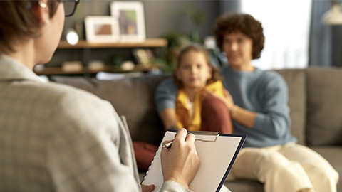 Close-up of child psychologist making notes in document while talking to mom and her son during her visit to home