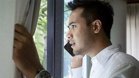 Concept: reporting a crime. Young Thai man standing in front of a window, looking outside and talking on his cellphone