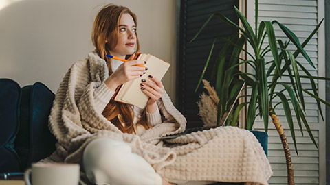 Thoughtful and cozy red-haired woman sitting with her legs crossed and taking notes on notebook